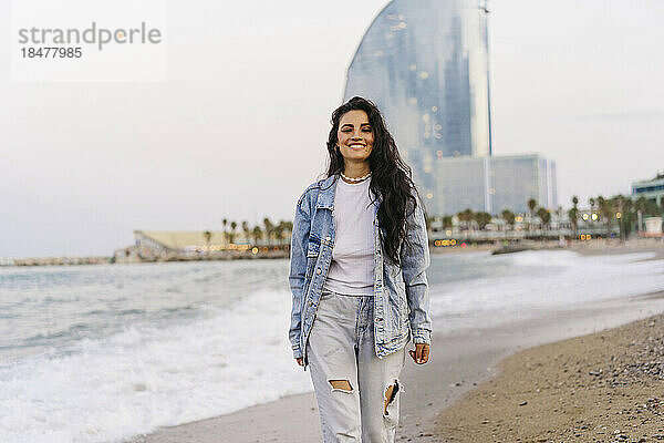 Glückliche Frau in Jeansjacke  die bei Sonnenuntergang am Strand spaziert