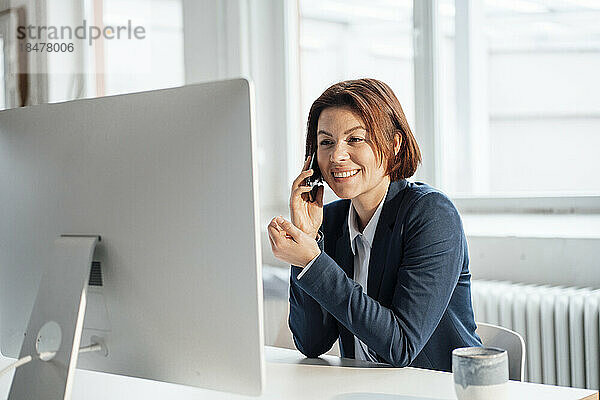 Glückliche Geschäftsfrau  die am Arbeitsplatz vor dem Desktop mit dem Smartphone spricht
