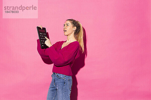 Nachdenkliche Frau mit Taschenrechner vor rosa Hintergrund