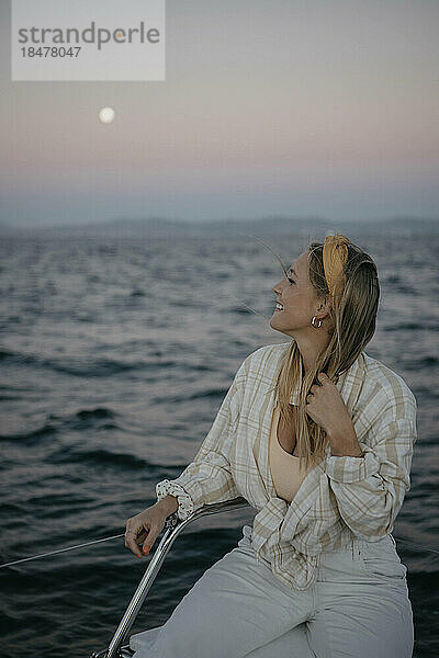 Lächelnde Frau sitzt bei Sonnenuntergang auf einem Boot im Meer