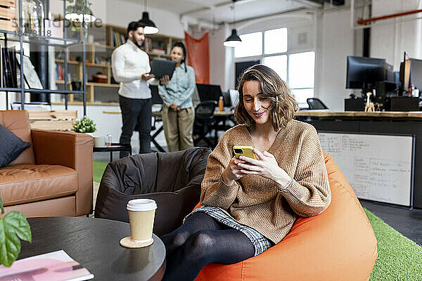 Lächelnde Geschäftsfrau  die im Büro auf einem Sitzsack sitzt und ihr Mobiltelefon benutzt