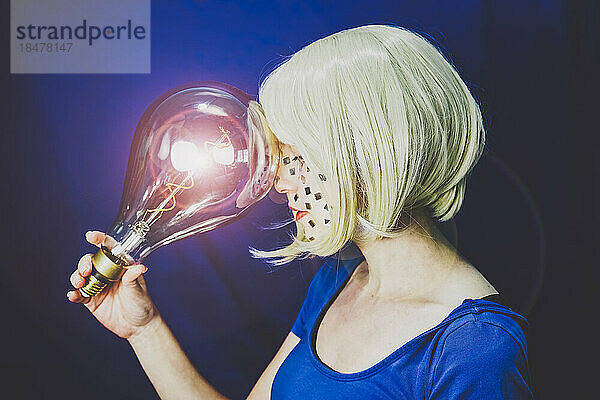 Blonde Frau hält große beleuchtete Glühbirne vor blauem Hintergrund