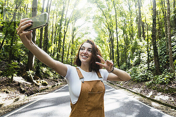 Glückliche junge Frau zeigt eine Geste des Friedenszeichens und macht ein Selfie vor Bäumen