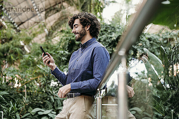Lächelnder Geschäftsmann benutzt Smartphone am Geländer vor Pflanzen