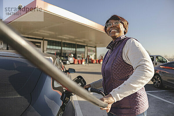 Glückliche ältere Frau  die an einem sonnigen Tag ein Elektroauto auflädt