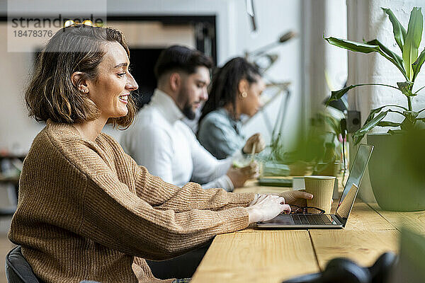Lächelnde Geschäftsfrau mit Einwegbecher sitzt mit Laptop im Büro