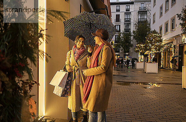 Lächelnde junge Frau hält Regenschirm in der Hand und steht mit Freundin auf Fußweg