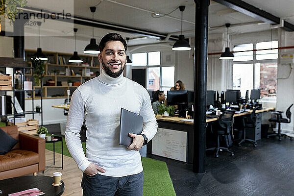 Lächelnder junger Geschäftsmann steht mit Laptop im Büro