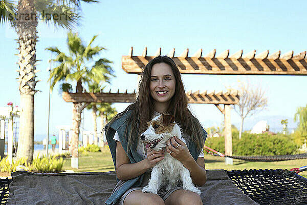 Glückliche junge Frau mit Hund sitzt auf Hängematte im Park