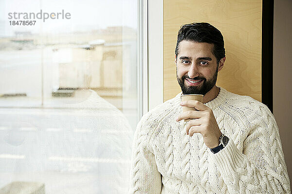 Lächelnder Mann im Pullover hält Kaffeetasse am Fenster
