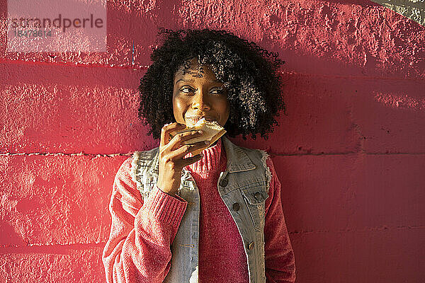 Lächelnde junge Frau mit Sandwich  die vor der roten Wand steht