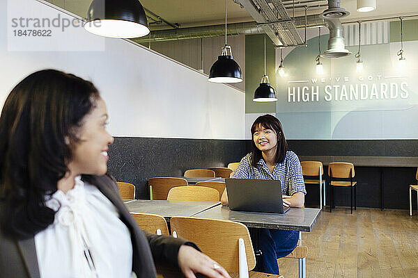 Lächelnde Geschäftsfrau im Gespräch mit einem Kollegen  der in der Cafeteria des Büros sitzt