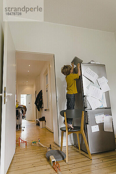 Boy reaching for book standing on chair near refrigerator at home