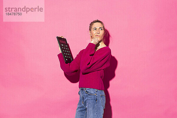 Verwirrte Frau mit Taschenrechner vor rosa Hintergrund