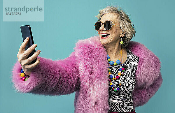 Glückliche Seniorin mit Schmuck macht ein Selfie mit dem Smartphone vor türkisfarbenem Hintergrund