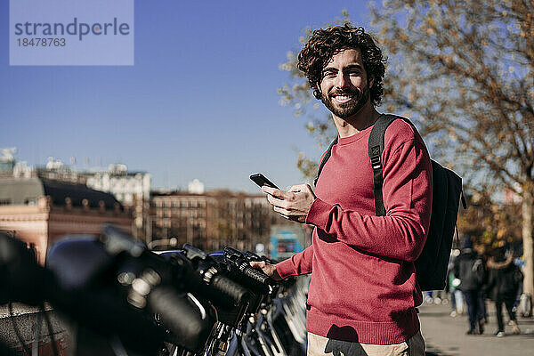 Lächelnder junger Mann mit Smartphone steht am Elektrofahrrad-Sharing-System