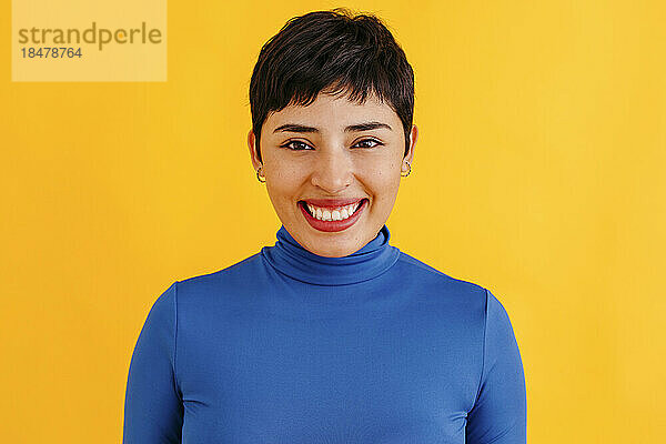 Glückliche junge Frau mit Pixie-Haarschnitt  die vor gelbem Hintergrund steht
