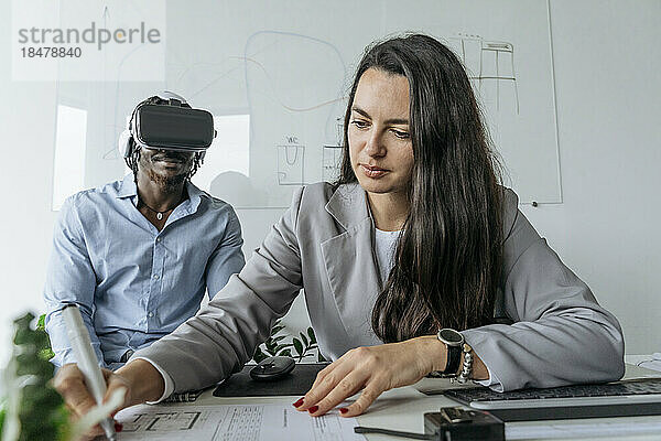Architekt bereitet Bauplan mit Kollege vor  der im Büro ein Virtual-Reality-Headset trägt