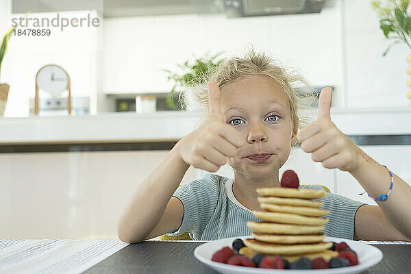 Girl making thumbs up gesture by plate of fresh pancakes at home