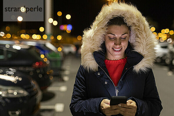 Glückliche Frau mit Parka-Kapuze  die nachts ihr Smartphone benutzt