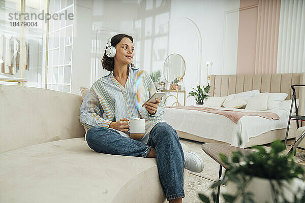 Lächelnde junge Frau mit kabellosen Kopfhörern sitzt zu Hause mit einer Kaffeetasse auf dem Sofa