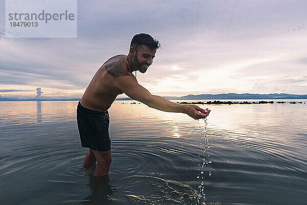 Glücklicher Mann  der mit Wasser spielt und den Sonnenuntergang am Strand genießt