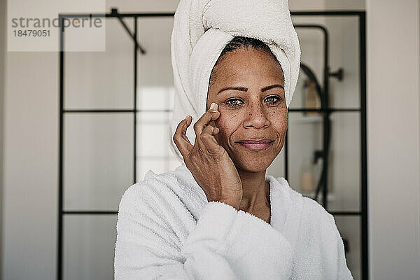 Lächelnde Frau  die zu Hause im Badezimmer Feuchtigkeitscreme auf ihr Gesicht aufträgt