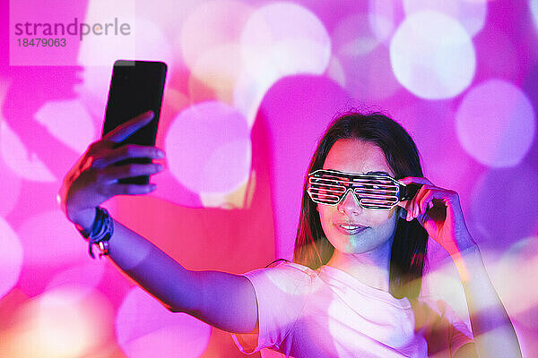 Junge Frau mit neuartiger Brille macht ein Selfie mit ihrem Smartphone in rosa Neonlichtern
