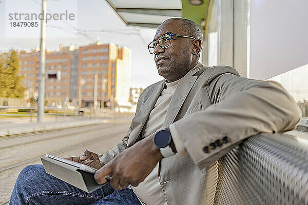 Nachdenklicher Geschäftsmann mit Tablet-PC sitzt an der Straßenbahnhaltestelle