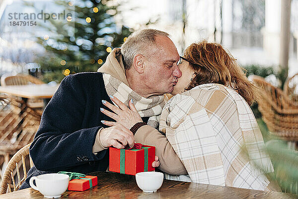Älterer Mann mit Geschenkbox küsst ältere Frau im Café