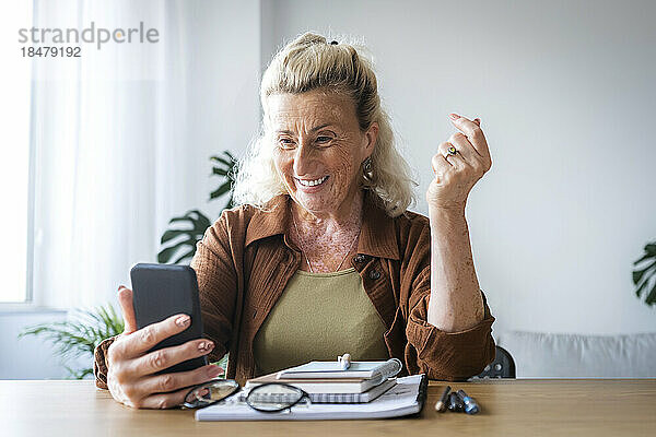 Glückliche ältere Geschäftsfrau hält Mobiltelefon am Schreibtisch im Heimbüro