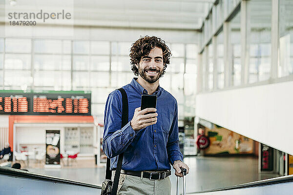 Lächelnder Geschäftsmann mit Mobiltelefon steht am Bahnhof