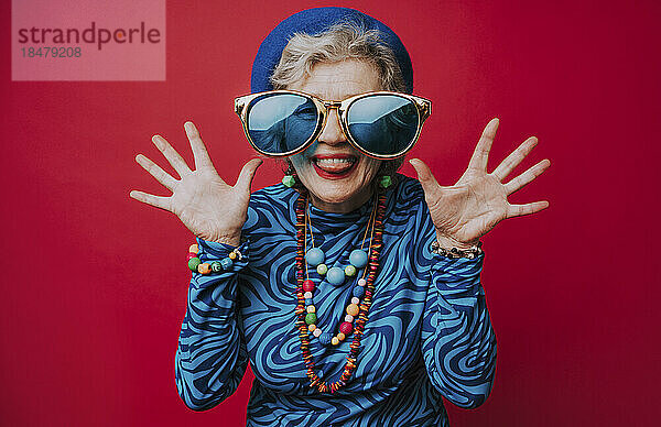 Ältere Frau mit großer Sonnenbrille amüsiert sich vor rotem Hintergrund