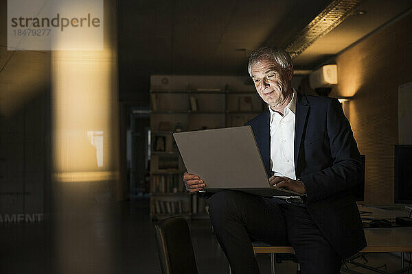 Lächelnder Geschäftsmann sitzt und arbeitet mit Laptop im Büro