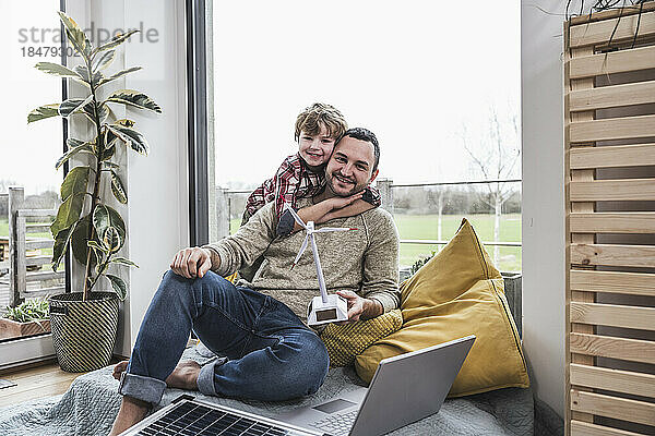 Glücklicher Vater hält Windturbinenmodell in der Hand und Sohn umarmt sich zu Hause von hinten