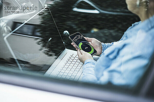Reife Frau mit Laptop und Lade-App für Elektroautos auf mobilem Bildschirm  durch Glas gesehen