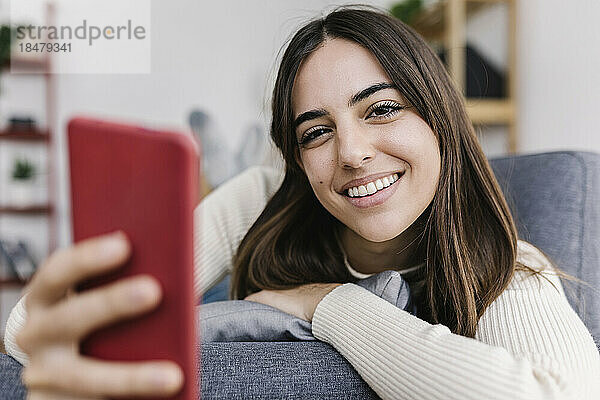 Lächelnde Frau mit Smartphone  die zu Hause auf dem Sofa liegt