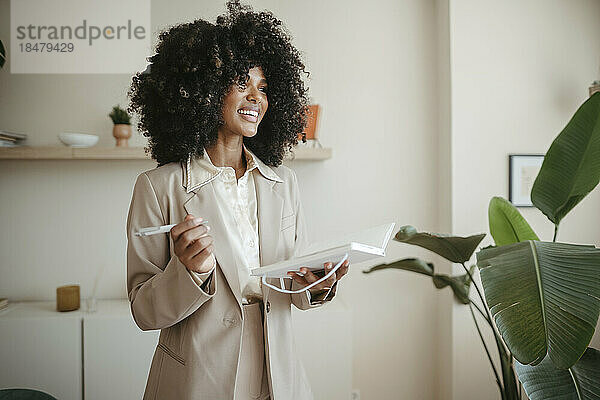 Glückliche Geschäftsfrau mit Afro-Frisur steht mit Buch im Büro