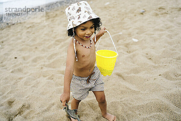 Netter Junge mit Hut  Eimer und Spielzeug im Sand am Strand