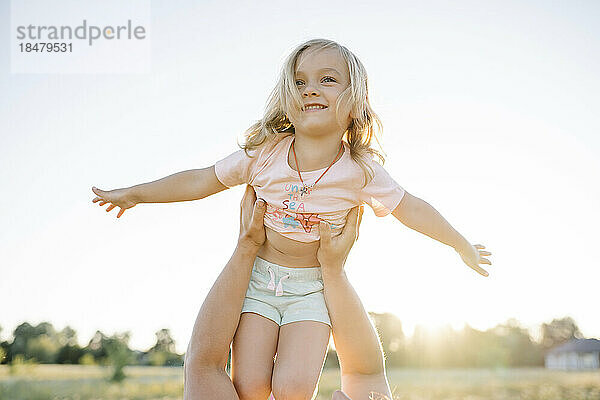 Mutter hebt an einem sonnigen Tag ein glückliches süßes Mädchen mit ausgestreckten Armen im Feld hoch