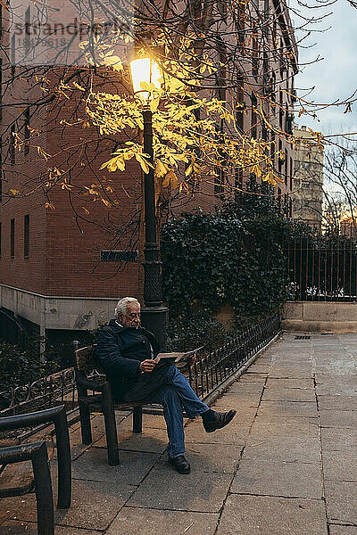 Älterer Mann liest ein Buch und sitzt auf einem Stuhl neben einem beleuchteten Laternenpfahl