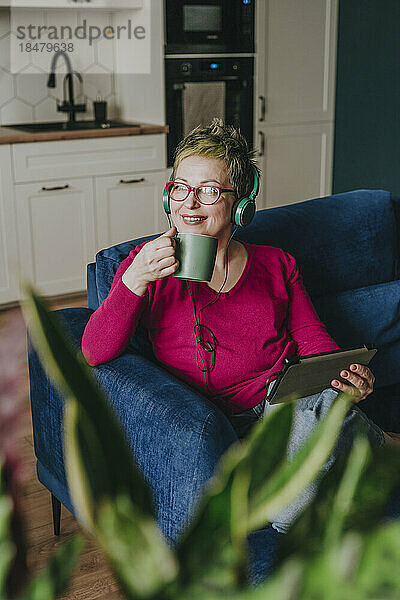 Thoughtful mature woman wearing headphones having coffee at home