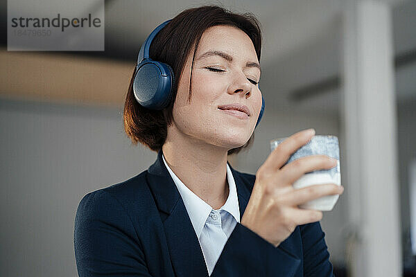 Junge Geschäftsfrau trägt Kopfhörer und hört Musik und hält eine Kaffeetasse im Büro