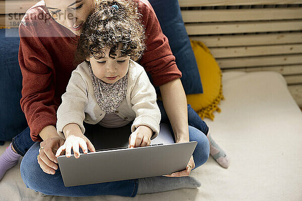 Kleines Mädchen sitzt mit Laptop auf dem Schoß der Mutter auf der Couch zu Hause