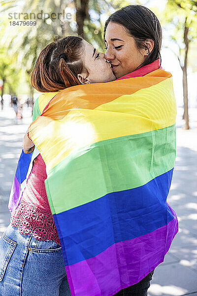 Lesbian couple covered in multi colored flag kissing on mouth at footpath