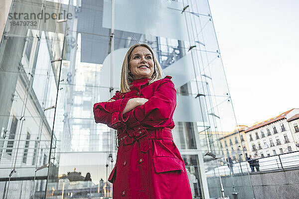 Lächelnde reife Frau mit verschränkten Armen steht vor einem modernen Glasgebäude