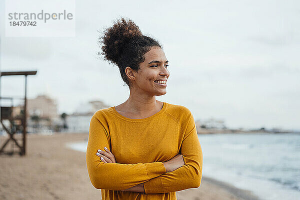Lächelnde junge Frau steht mit verschränkten Armen am Strand