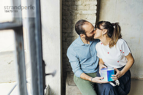 Paar mit Geschenkbox küsst sich am Fenster sitzend
