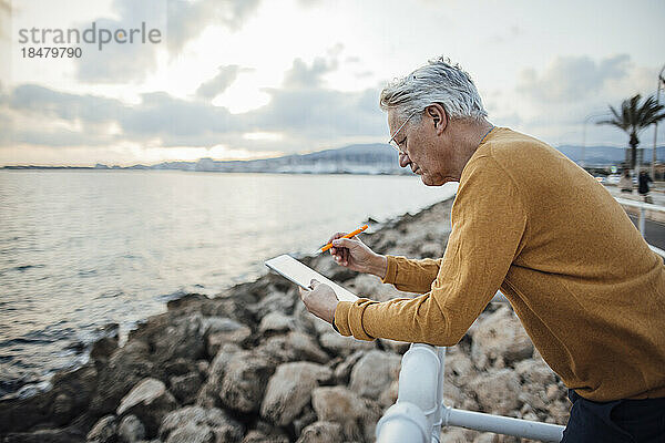 Älterer Mann benutzt Tablet-PC und lehnt am Geländer vor dem Meer