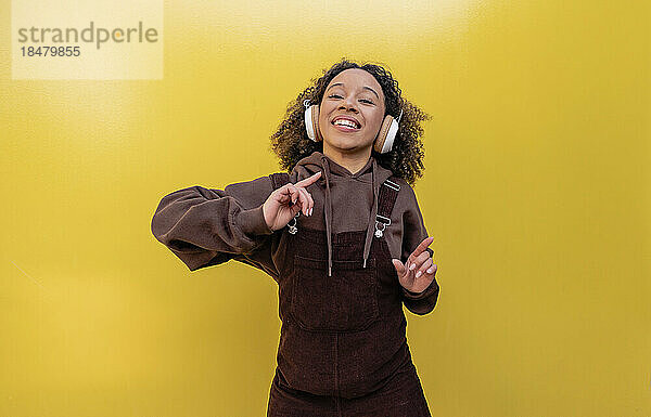 Glückliche Frau mit Kopfhörern tanzt vor gelber Wand